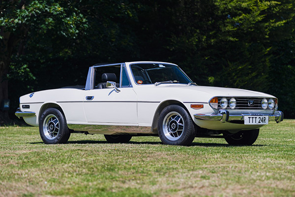 White Triumph Stag convertible on grass with trees backdrop