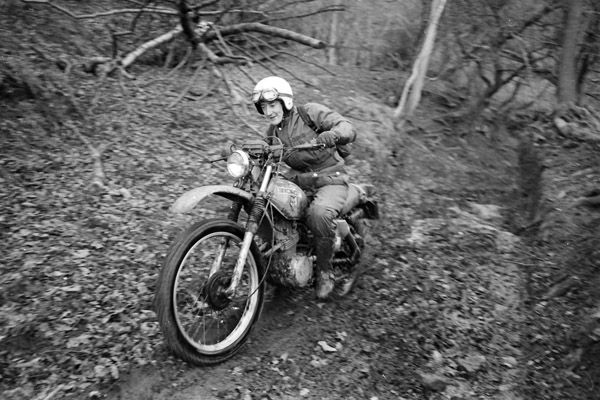 A black and white photo of a man riding a classic bike through woodland