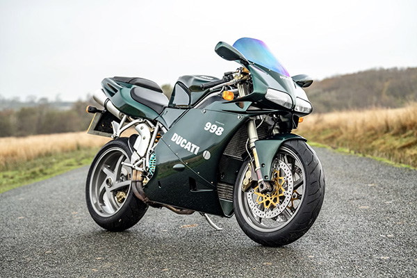 2004 Racing Green Ducati 998 Matrix Parked Sideways On A Road Surrounded By Grass And Hills With Trees