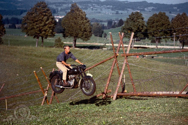 Steve Mcqueen In The Air On A Black Triumph Tr6 Motorbike Beside Wooden Barbed Wire Fencing And A Mountain Backdrop A Scene From The Great Escape Film