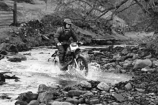 A black and white photo of a man riding a classic bike through a stream